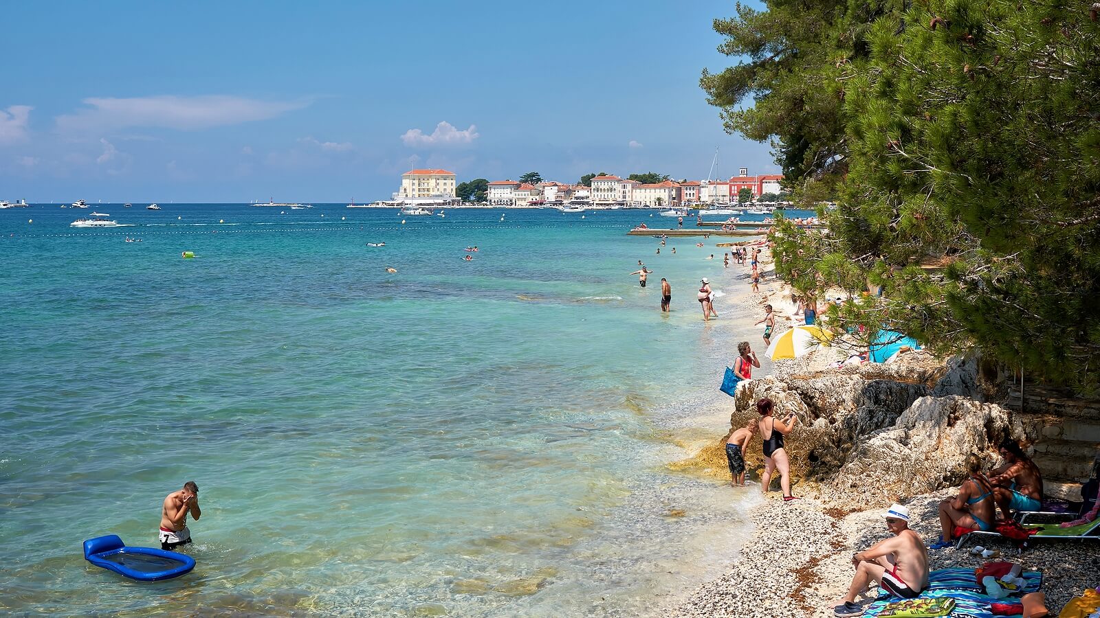 Crowded beach in Poreč, Croatia on a sunny summer day, with people swimming in the turquoise sea, sunbathing on the pebbled shore, and boats visible in the distance near the old town.