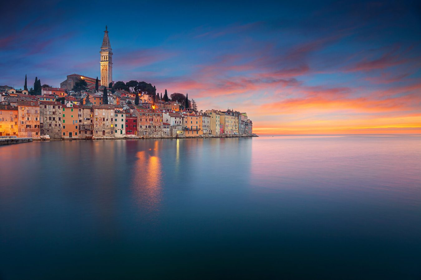Romantic sunset view of Rovinj, Croatia, with colorful historic buildings lining the waterfront and the prominent Church of St. Euphemia rising above the old town, reflecting in the calm Adriatic Sea.